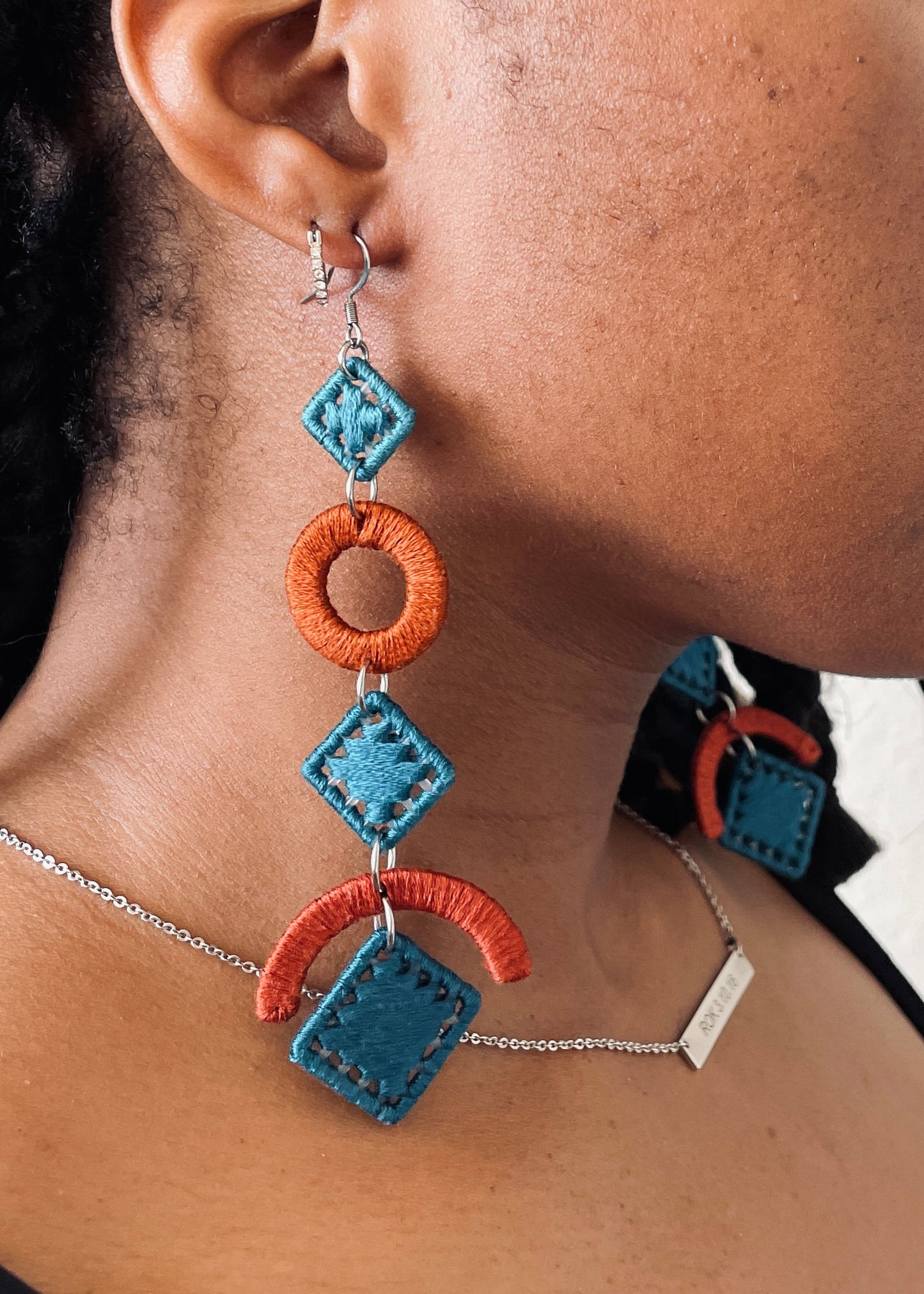 A pair of OlaNti handcrafted statement earrings featuring bold geometric shapes in vibrant orange and deep teal, displayed on a stand with soft shadows. A close-up of the earrings worn, showcasing their lightweight movement and intricate detailing