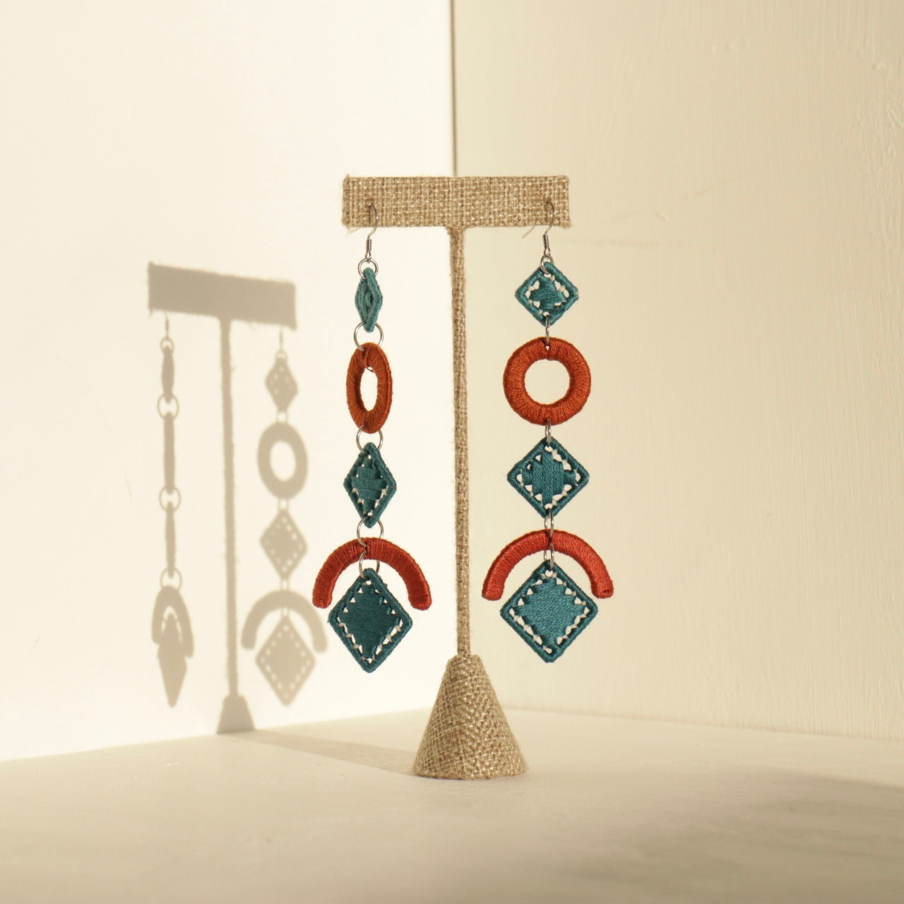A pair of handcrafted statement earrings featuring bold geometric shapes in dark orange and deep teal, displayed on a stand with soft shadows. A close-up of the earrings worn, showcasing their lightweight movement and intricate detailing