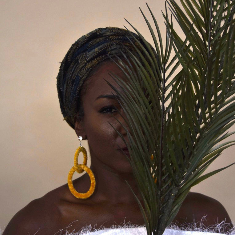 Model wearing The Valente earrings in yellow, partially concealed by palm leaves
