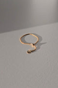 A delicate gold chain ring with a small dangling heart charm, displayed on a textured gray surface under warm, directional sunlight.