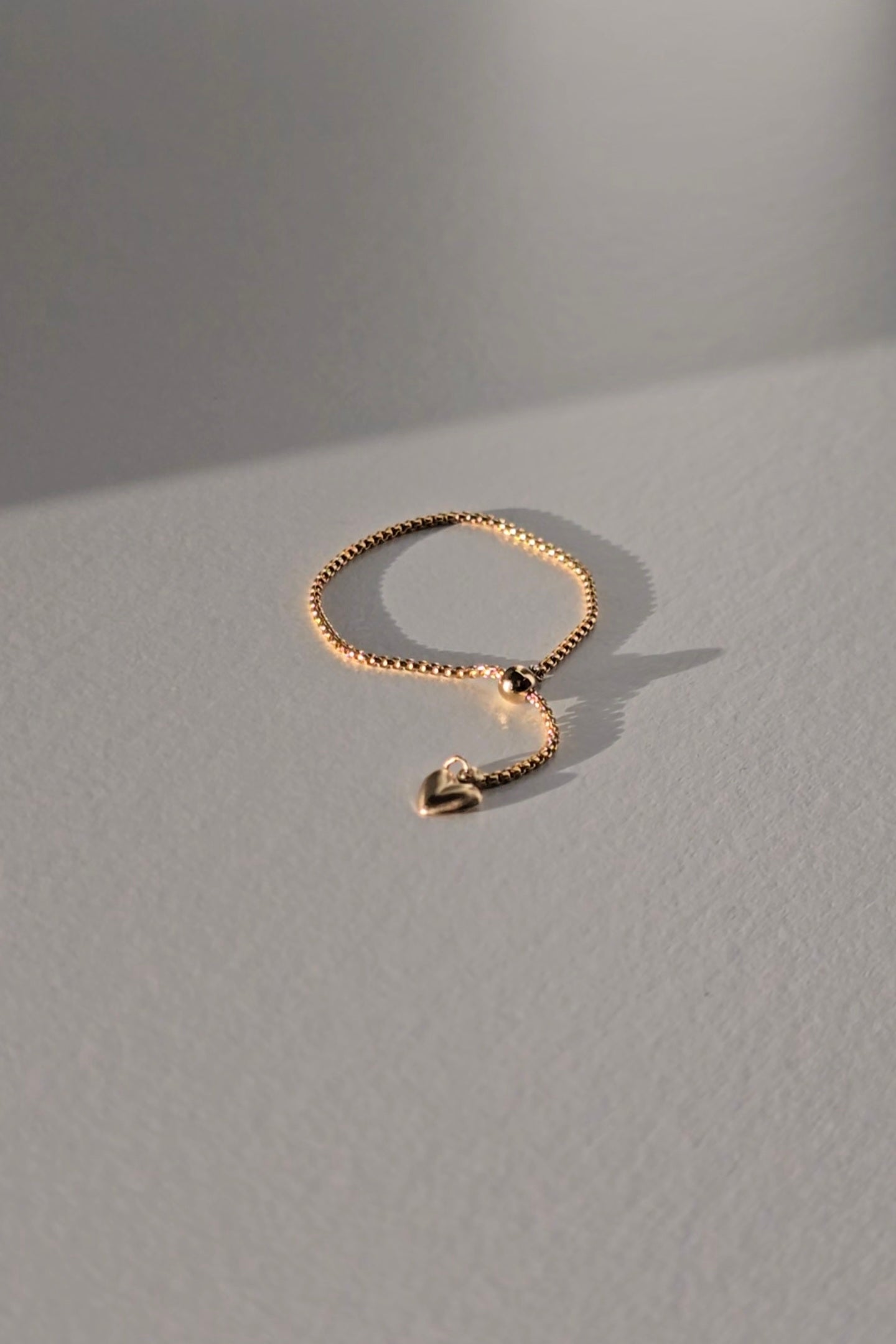 A delicate gold chain ring with a small dangling heart charm, displayed on a textured gray surface under warm, directional sunlight.