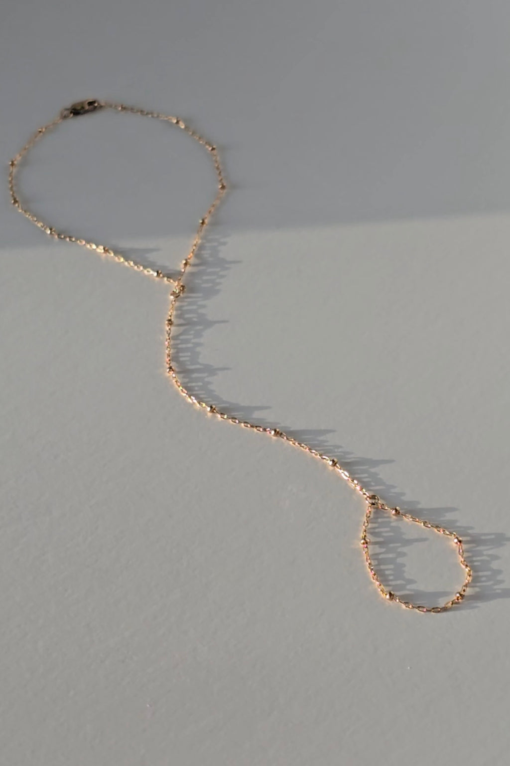 A delicate gold hand chain with textured, hammered links, laid out in a loose loop on a neutral gray surface with soft shadows.