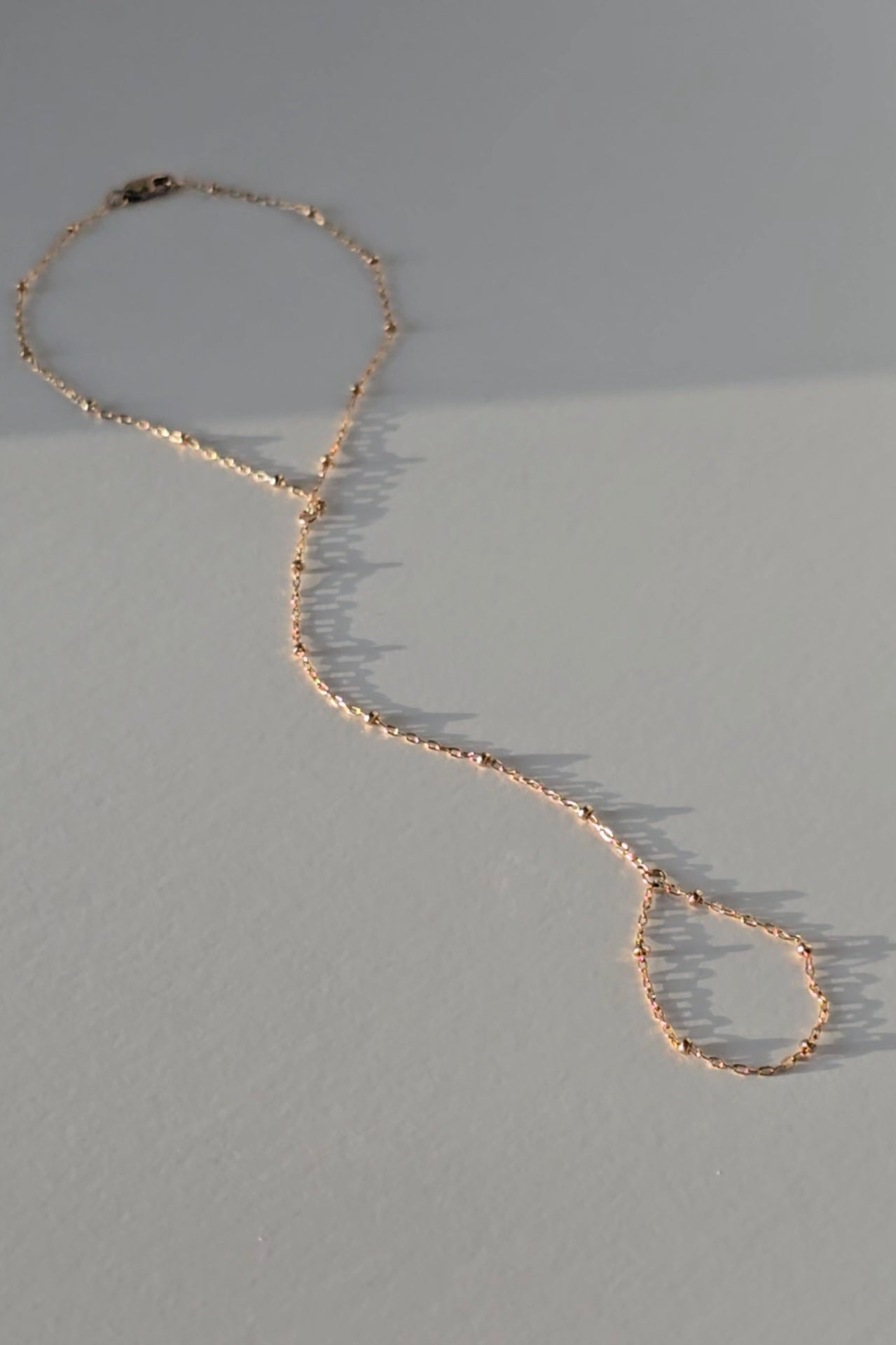 A delicate gold hand chain with textured, hammered links, laid out in a loose loop on a neutral gray surface with soft shadows.