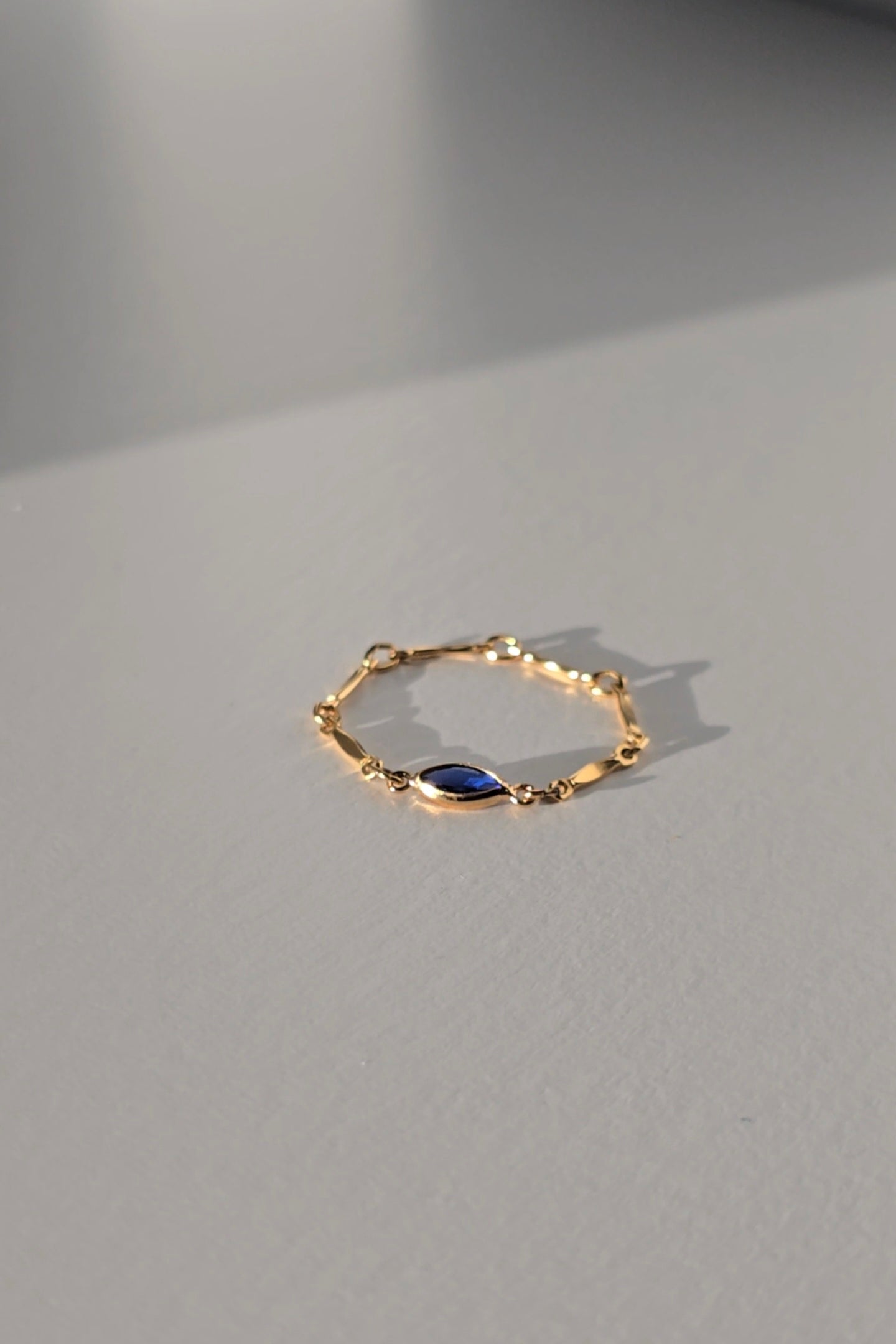 A delicate gold chain ring with a small marquise-cut dark blue stone in the center, resting on a light gray surface with soft shadows.