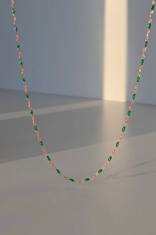 A delicate chain necklace featuring small green oval stones set in a rose gold-tone metal, suspended against a minimalist gray background with soft diagonal light beams.