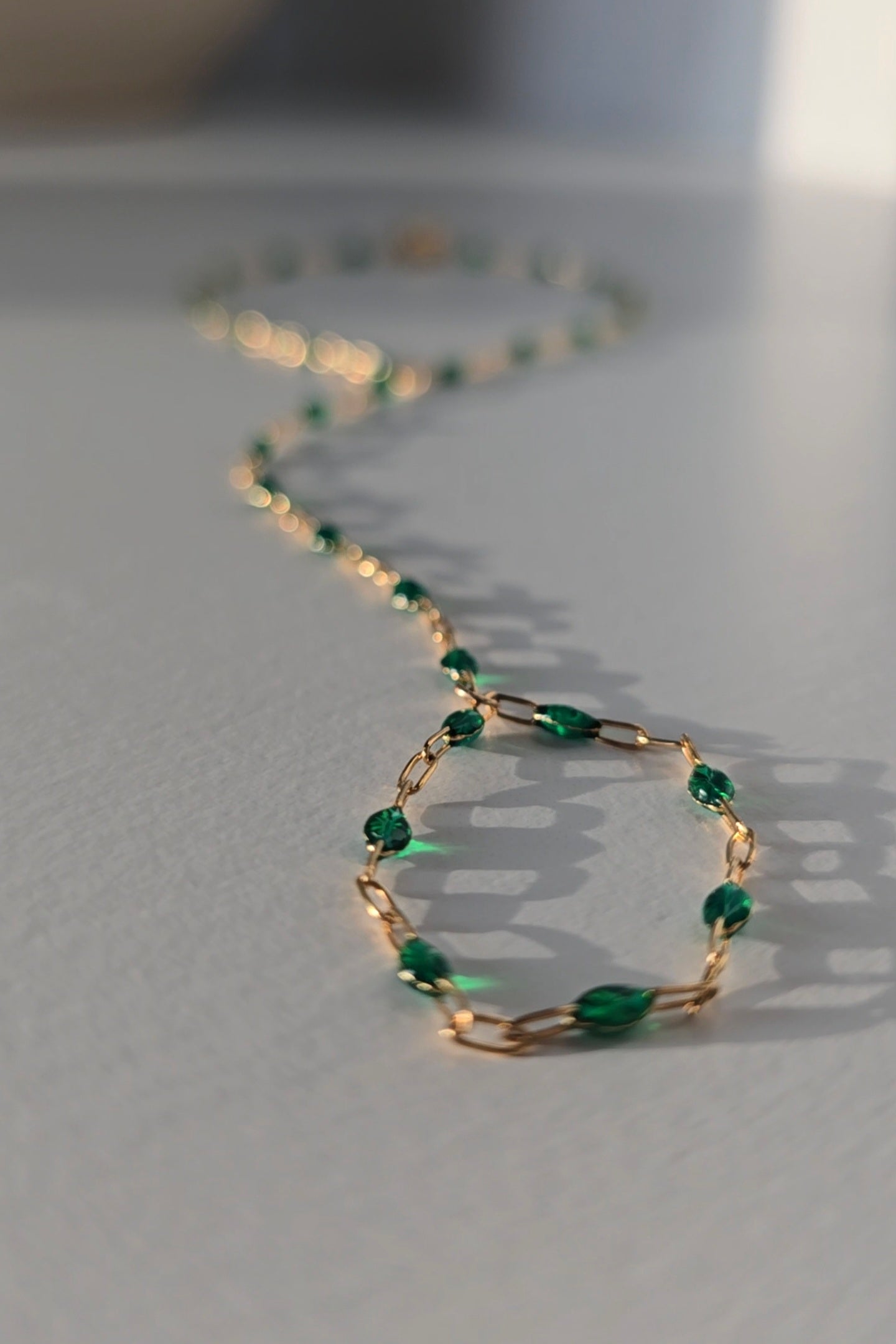 A delicate gold hand chain adorned with small, faceted green beads, arranged in a loose circle on a light gray surface with soft, natural shadows.