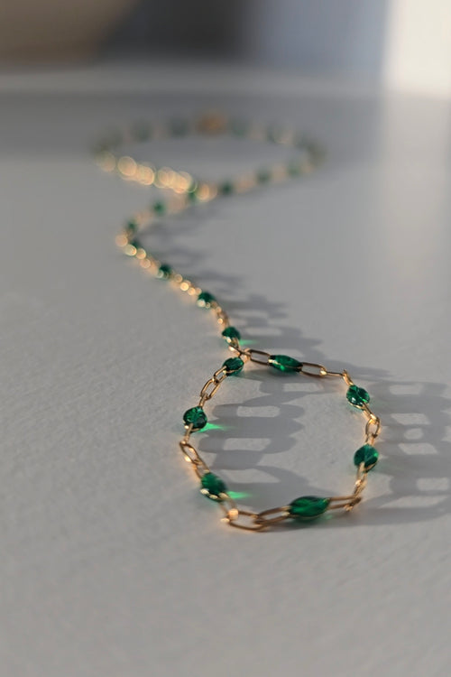 A delicate gold hand chain adorned with small, faceted green beads, arranged in a loose circle on a light gray surface with soft, natural shadows.