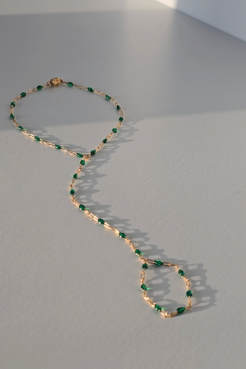 A delicate gold hand chain nwith small green faceted beads, arranged in a loop on a light gray surface with soft shadows.