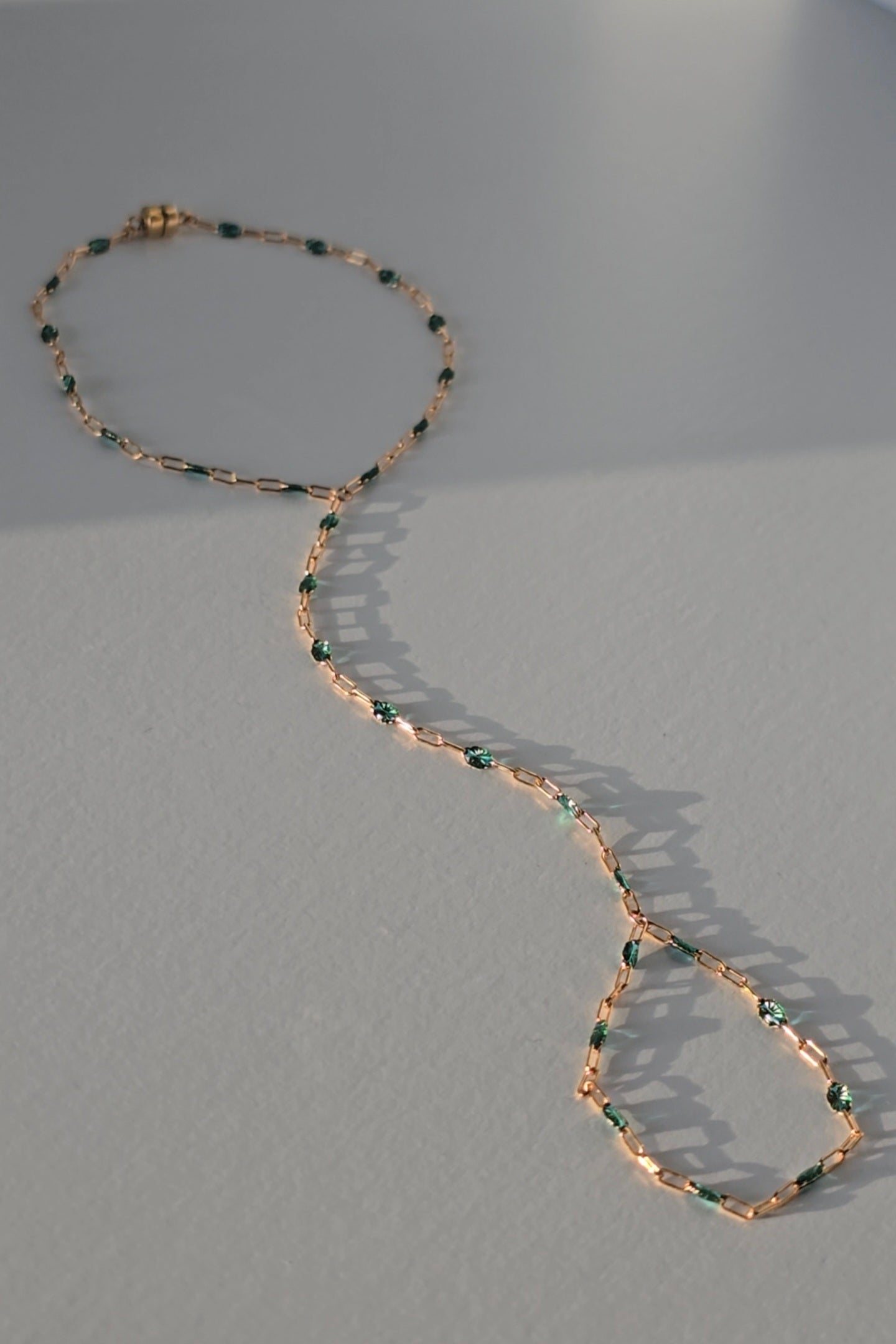 A delicate gold hand chain with small teal bead accents, arranged in a loose loop on a light gray surface with soft shadows.