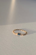 A delicate gold chain ring featuring a small rectangular blue gemstone, resting on a textured white surface with soft, warm sunlight and shadows.