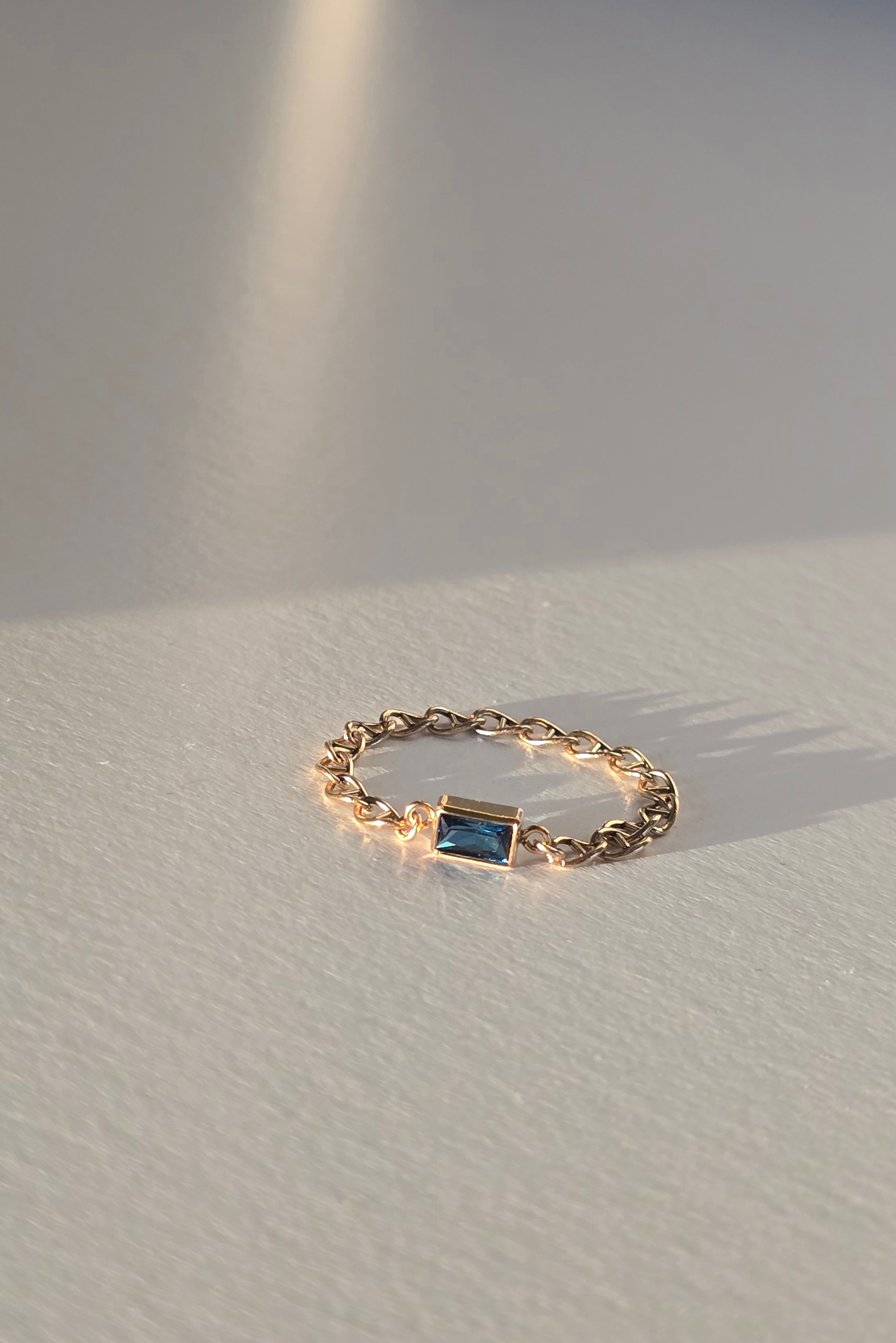 A delicate gold chain ring featuring a small rectangular blue gemstone, resting on a textured white surface with soft, warm sunlight and shadows.