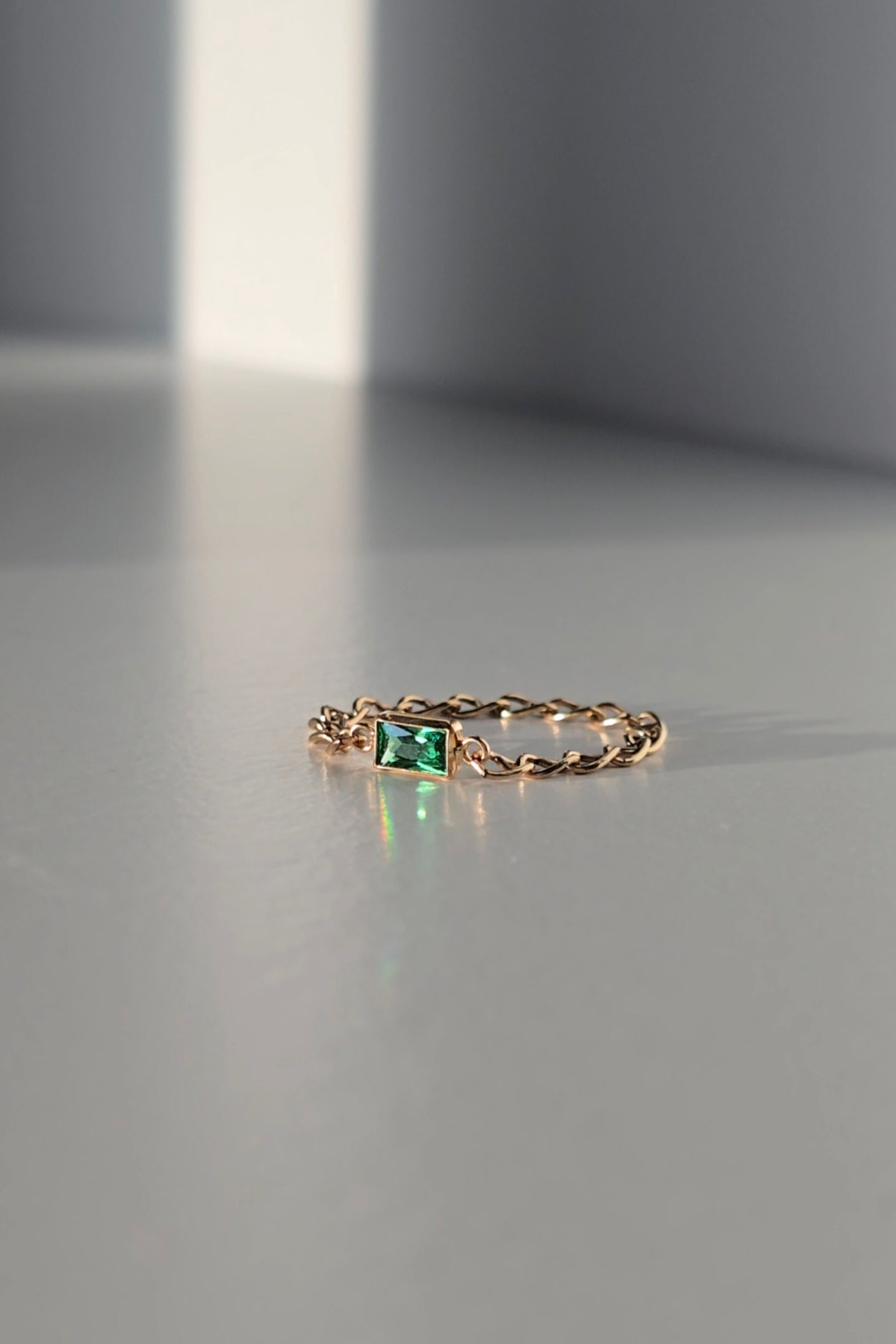 A delicate gold chain ring featuring a small rectangular green emerald-cut gemstone in the center, resting on a minimalist gray surface with soft shadows.