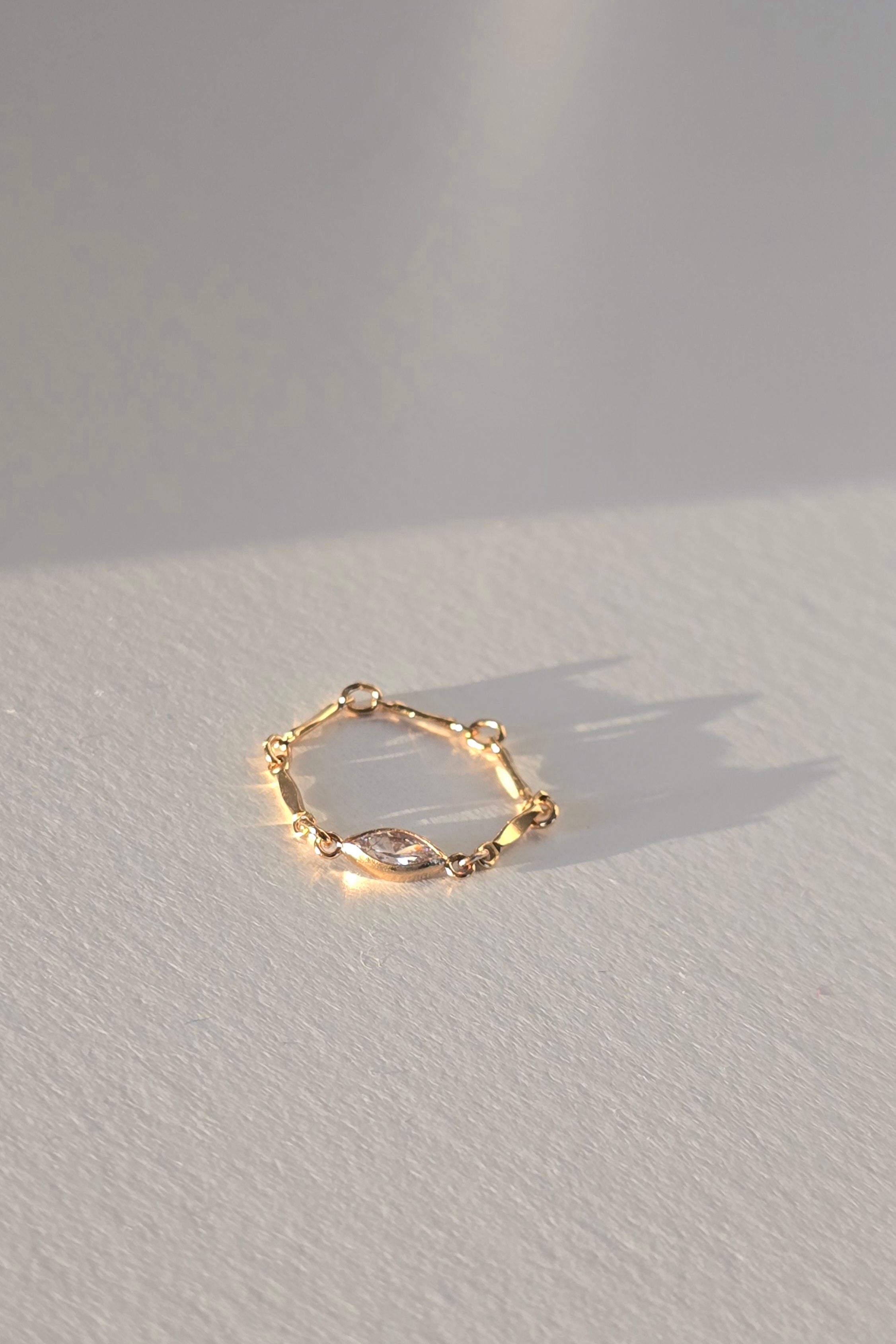 A delicate gold ring with a marquise-cut clear gemstone, resting on a textured white surface under warm, directional sunlight that casts a soft shadow.