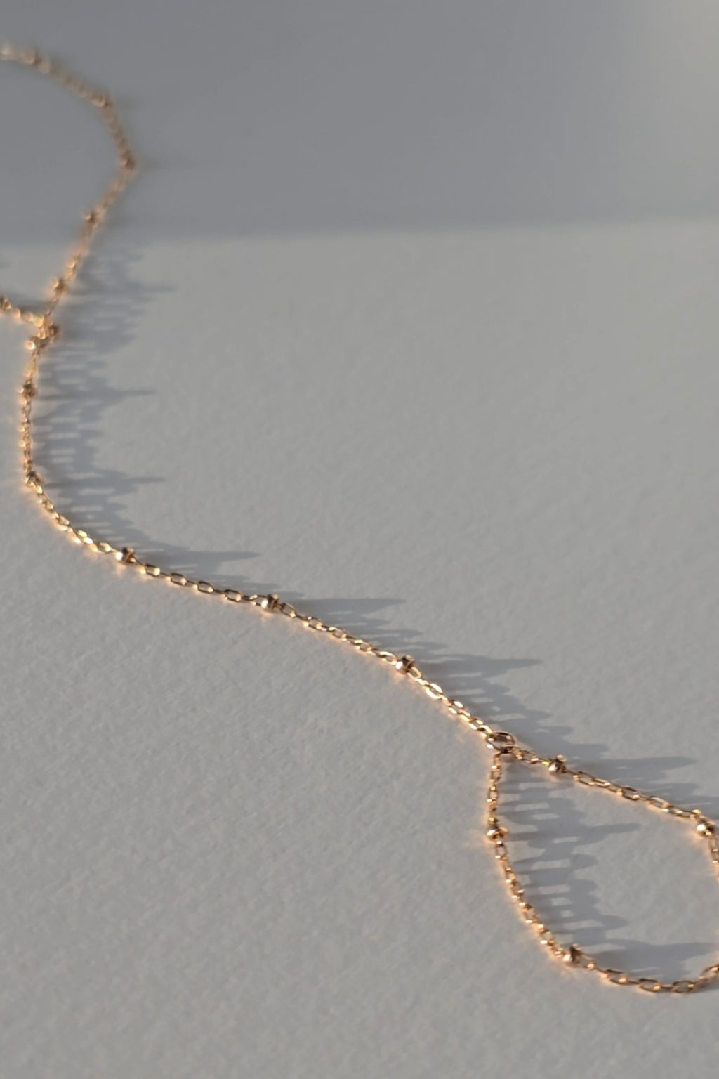 A delicate gold hand chain draped diagonally across a textured light gray surface, casting soft shadows.