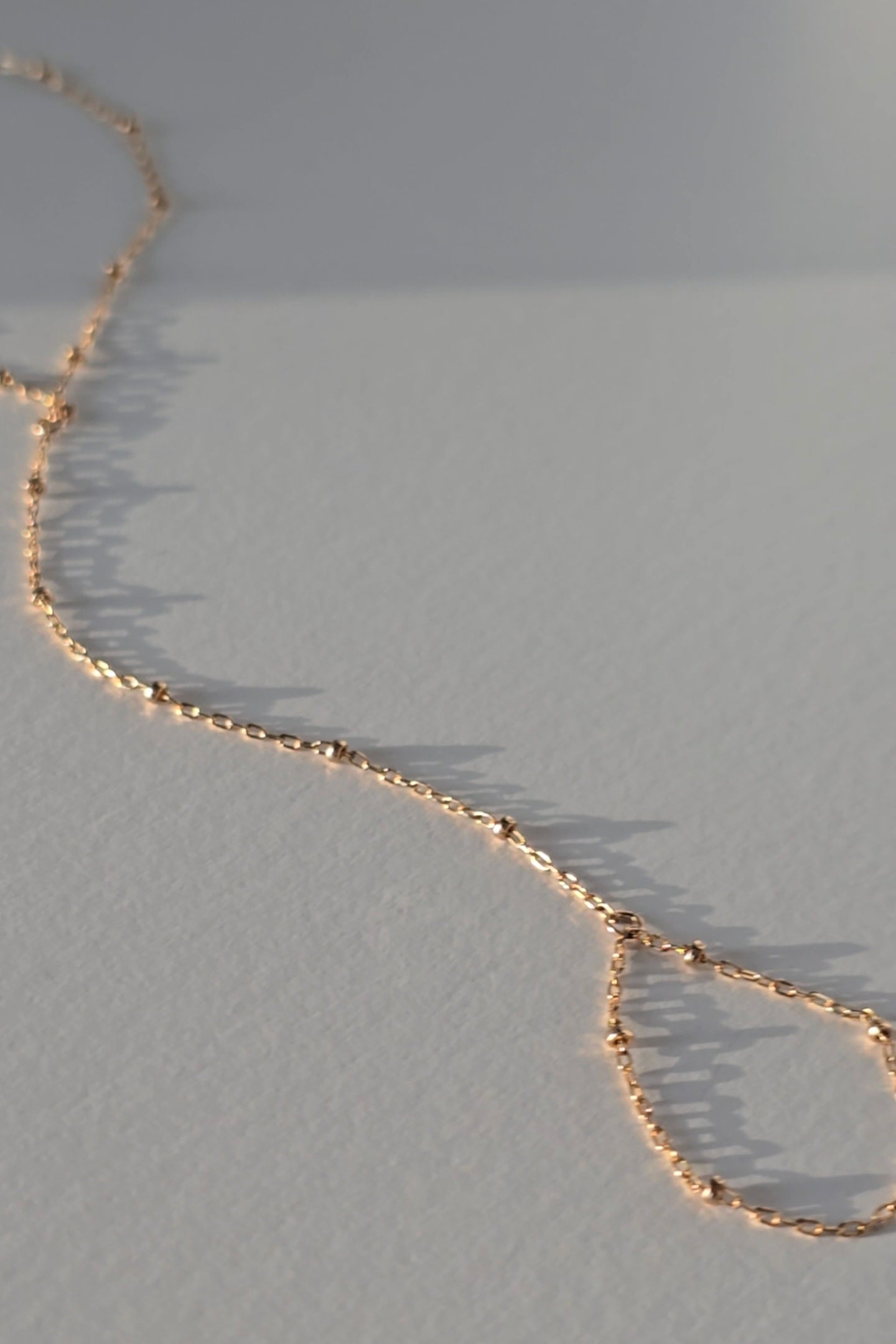 A delicate gold hand chain draped diagonally across a textured light gray surface, casting soft shadows.