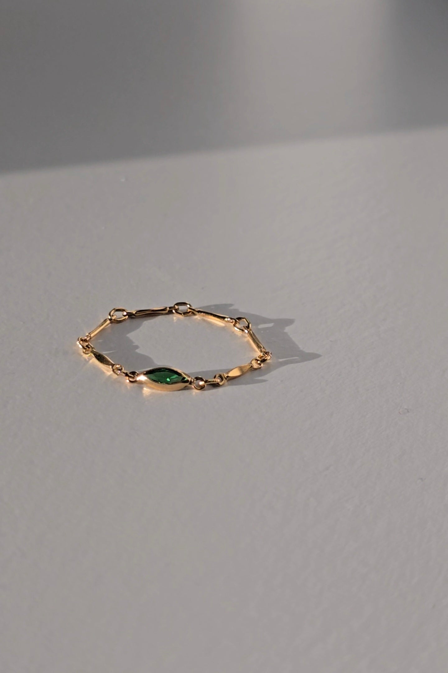 A delicate gold chain ring with a central marquise-cut green gemstone, resting on a neutral gray surface with soft shadows.