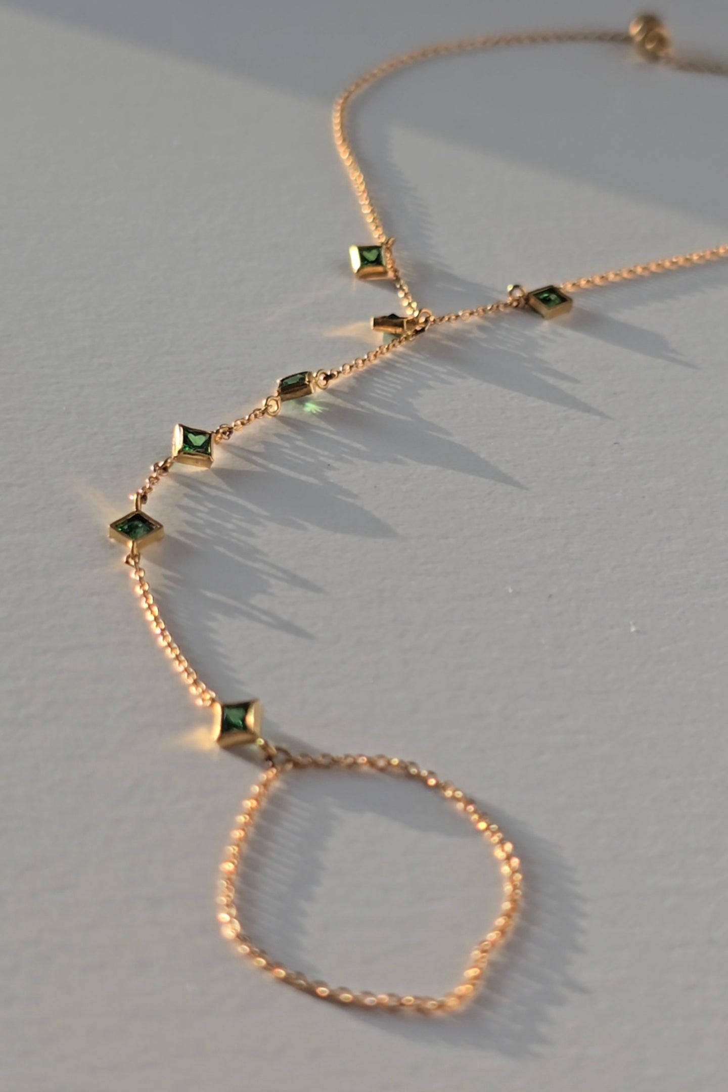 A delicate gold hand chain with small diamond-shaped gold pendants set with green stones, arranged on a light gray surface with soft shadows.