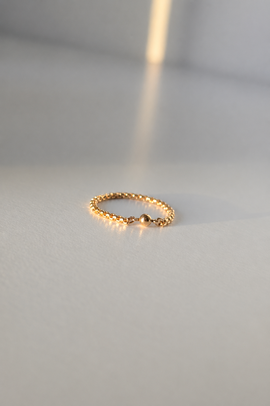 A delicate gold chain ring with a small central bead, resting on a light gray surface with a soft shadow.
