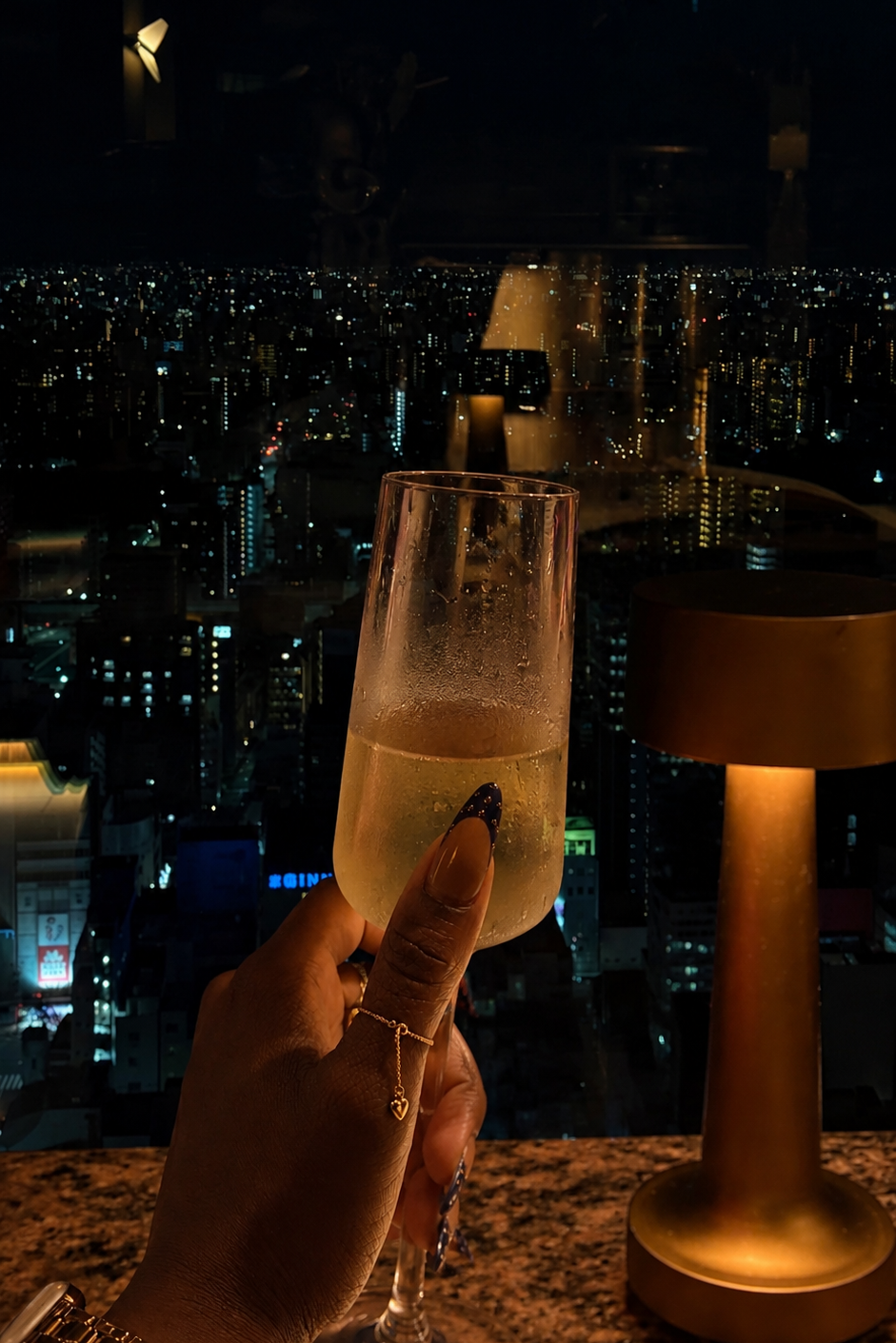Woman holding a glass to the city view at night wearing a gold chain ring
