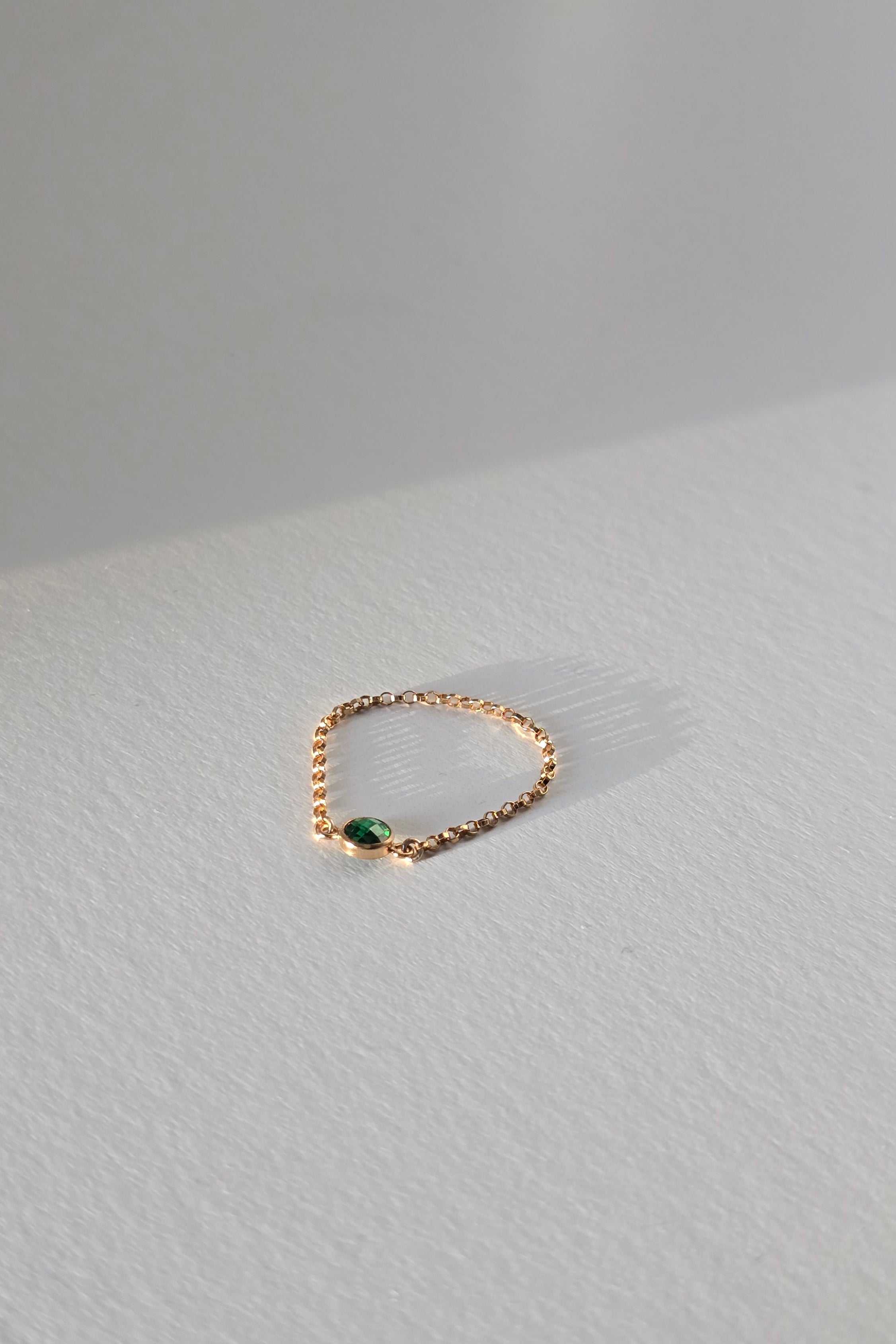 A delicate rose gold chain ring with a small green oval gemstone, resting on a textured light gray surface.