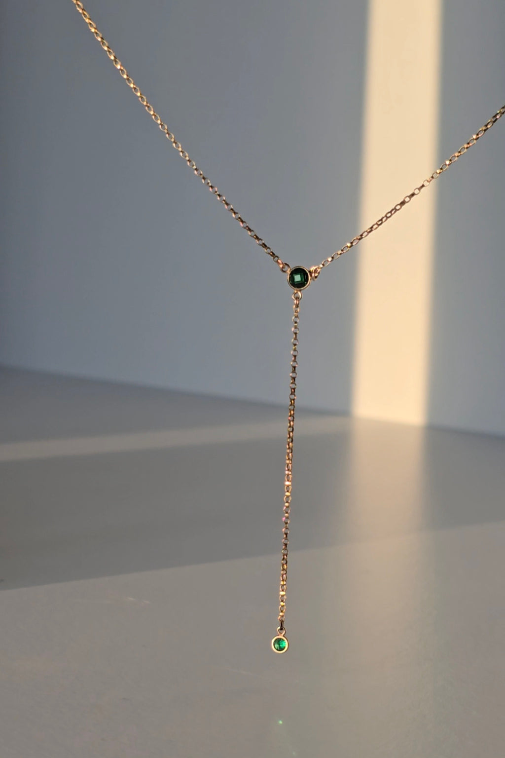 A delicate rose gold Y-necklace with a short drop chain, featuring a small dark green gemstone at the center and a larger green gemstone at the bottom, displayed against a minimalist gray background with warm sunlight casting soft shadows.