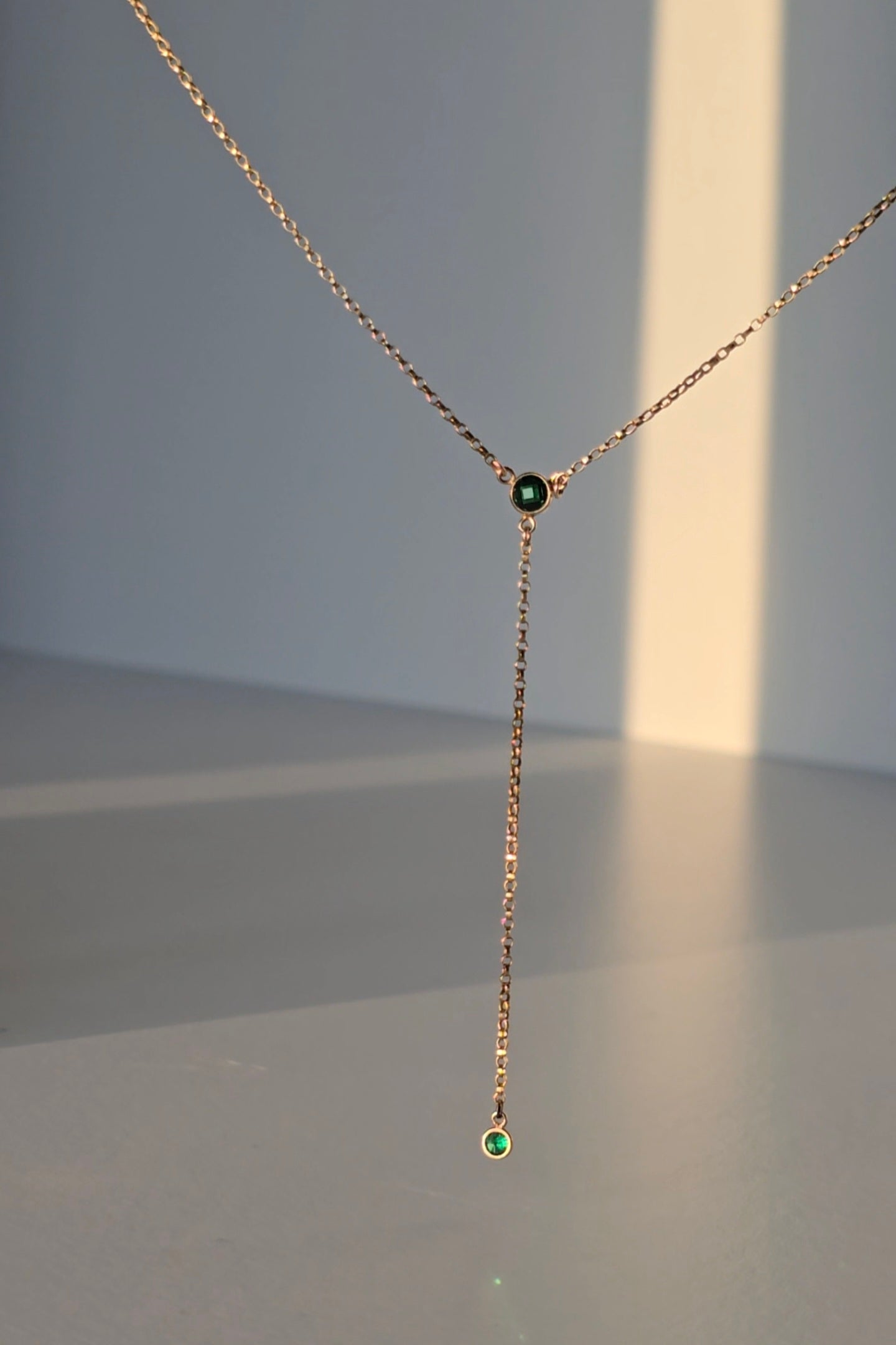 A delicate rose gold Y-necklace with a short drop chain, featuring a small dark green gemstone at the center and a larger green gemstone at the bottom, displayed against a minimalist gray background with warm sunlight casting soft shadows.