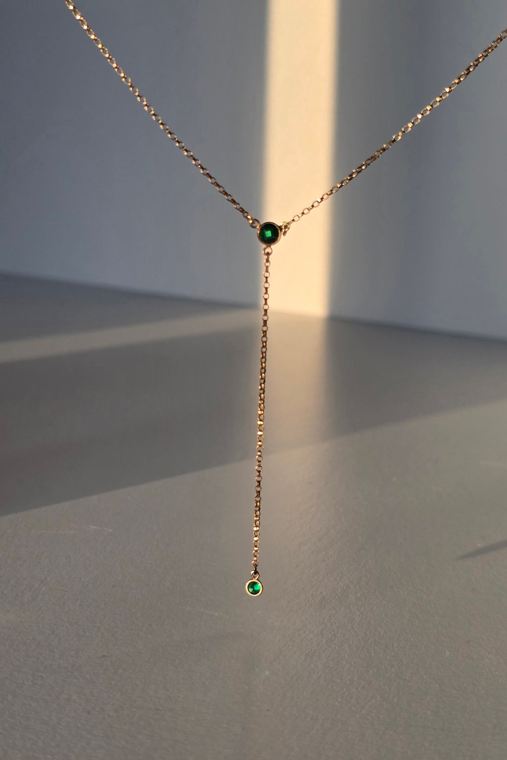 A delicate gold Y-necklace with a short drop chain and two small round green gemstone accents, displayed against a minimalist gray background with warm sunlight casting soft shadows.