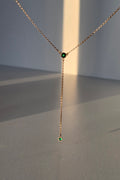 A delicate gold Y-necklace with a short drop chain and two small round green gemstone accents, displayed against a minimalist gray background with warm sunlight casting soft shadows.