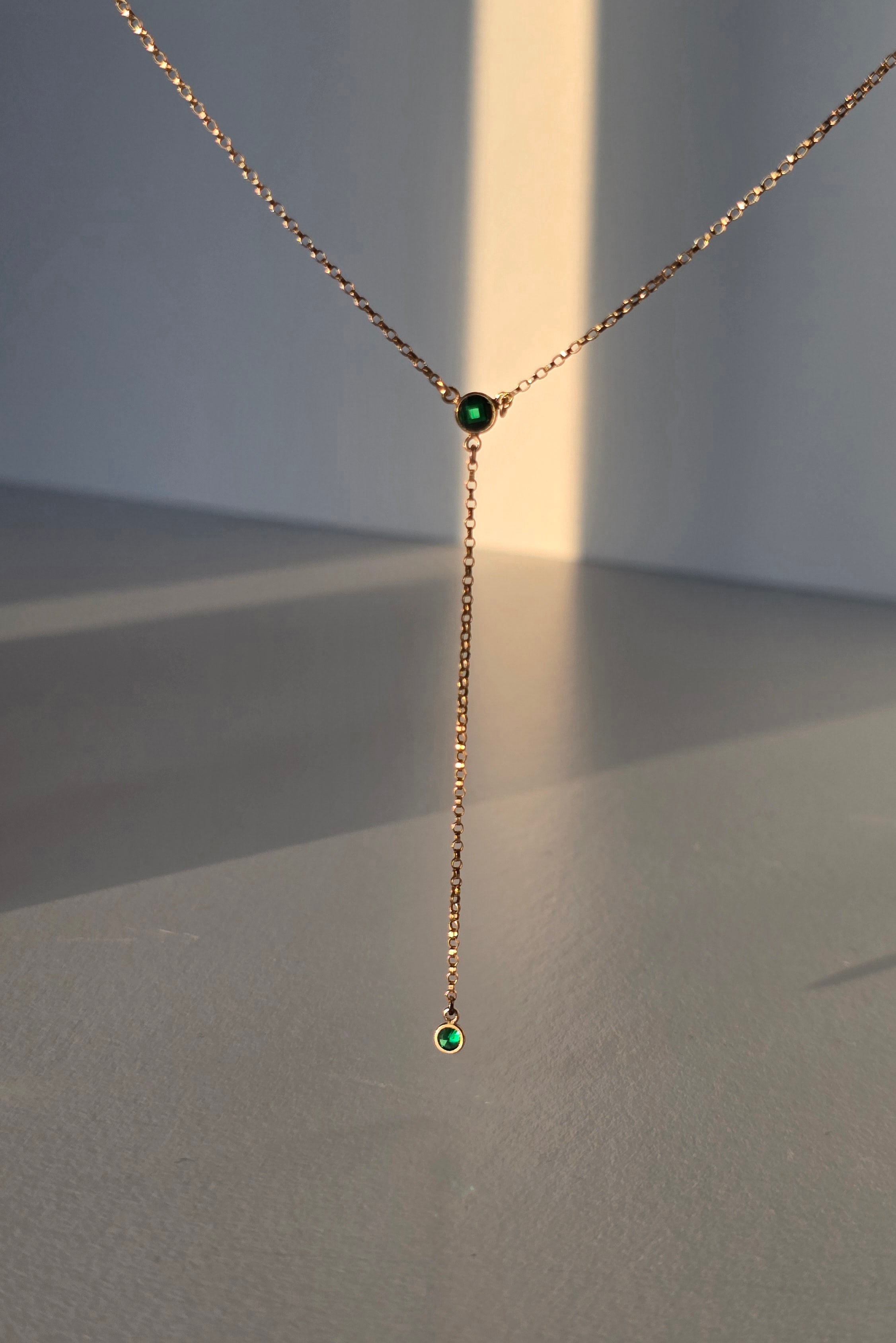 A delicate gold Y-necklace with a short drop chain and two small round green gemstone accents, displayed against a minimalist gray background with warm sunlight casting soft shadows.