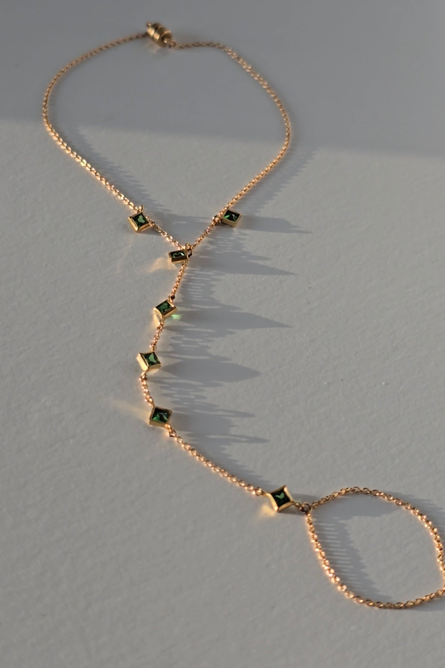 A gold hand chain with emeralds on a warm gray surface
