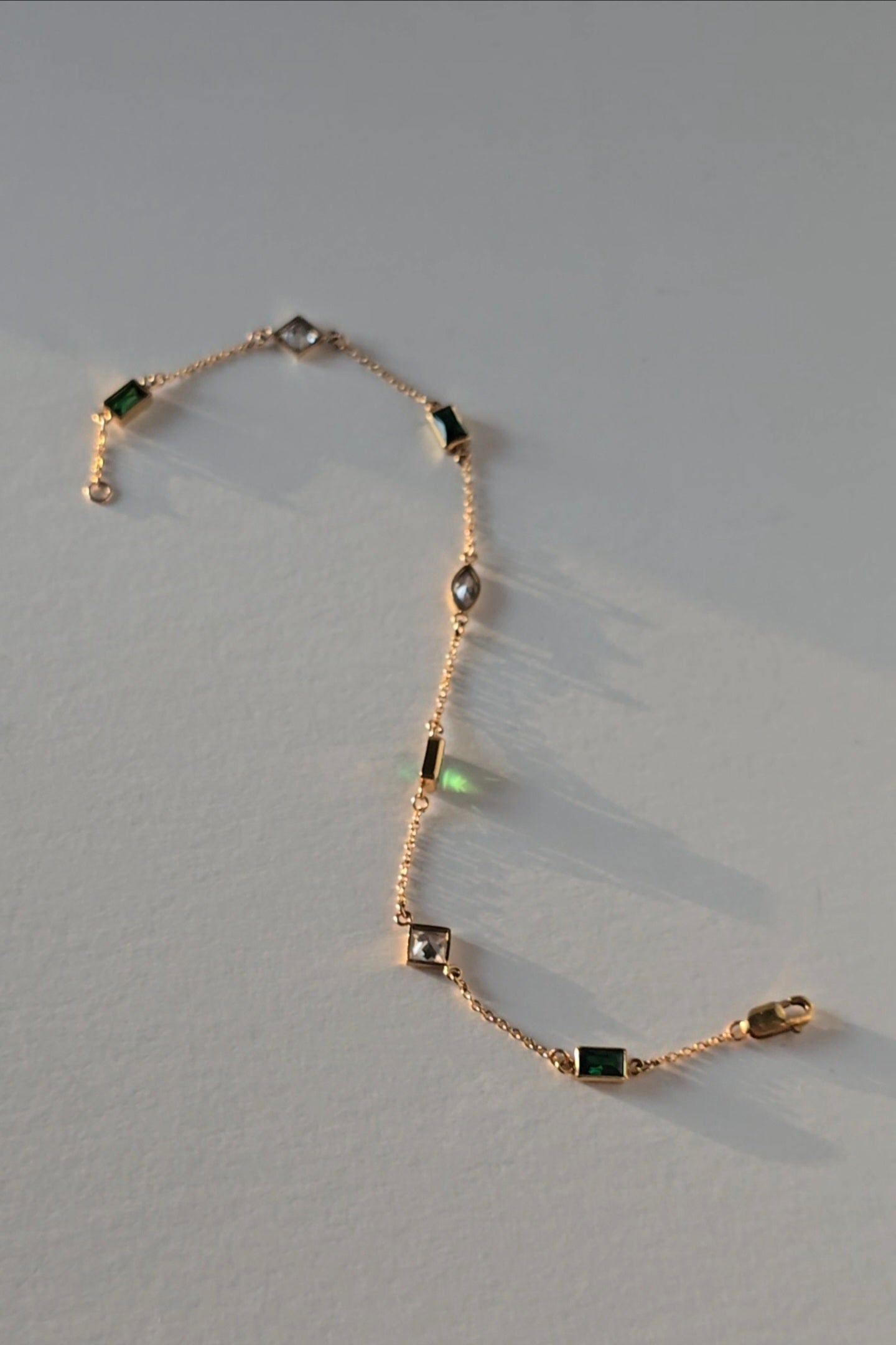 A delicate gold chain bracelet featuring alternating rectangular green emerald-cut stones and marquise-cut clear crystals, displayed on a neutral light gray background.