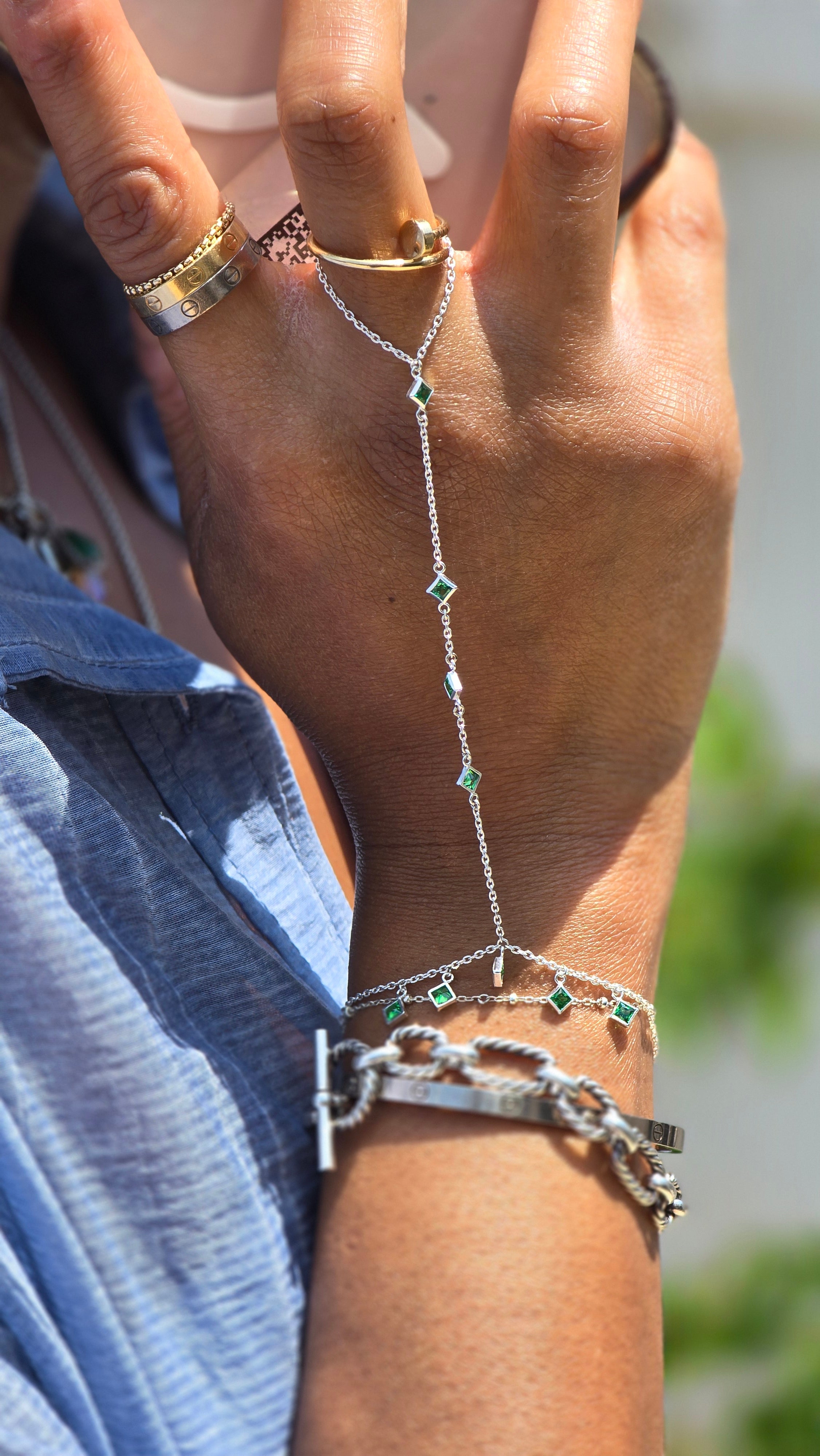 A close-up of a person's hand and wrist wearing a delicate silver chain hand bracelet with small green stone accents, along with other jewelry and a blue striped shirt.