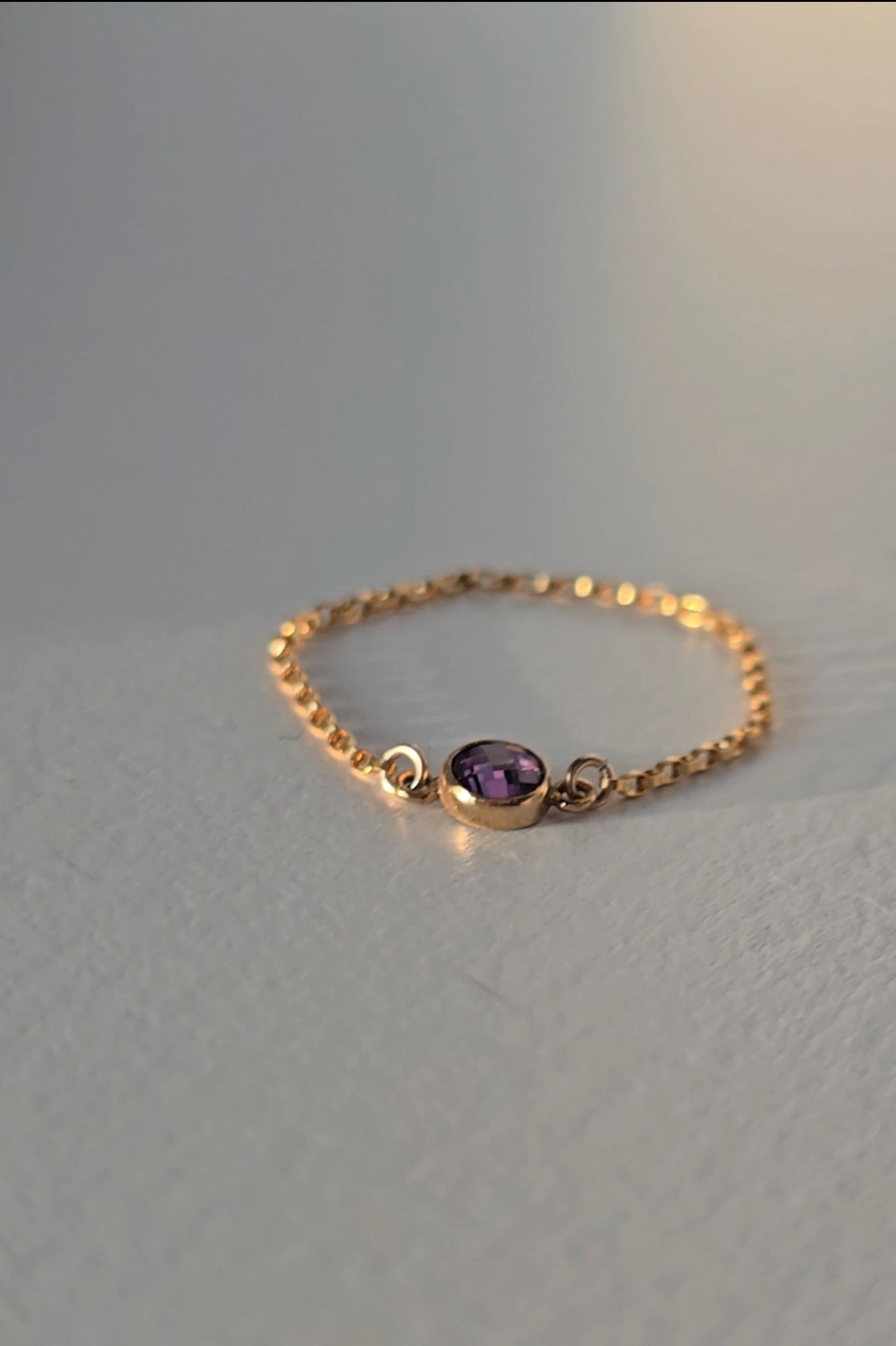 A delicate gold chain ring featuring a small oval purple gemstone, displayed on a neutral light gray surface.
