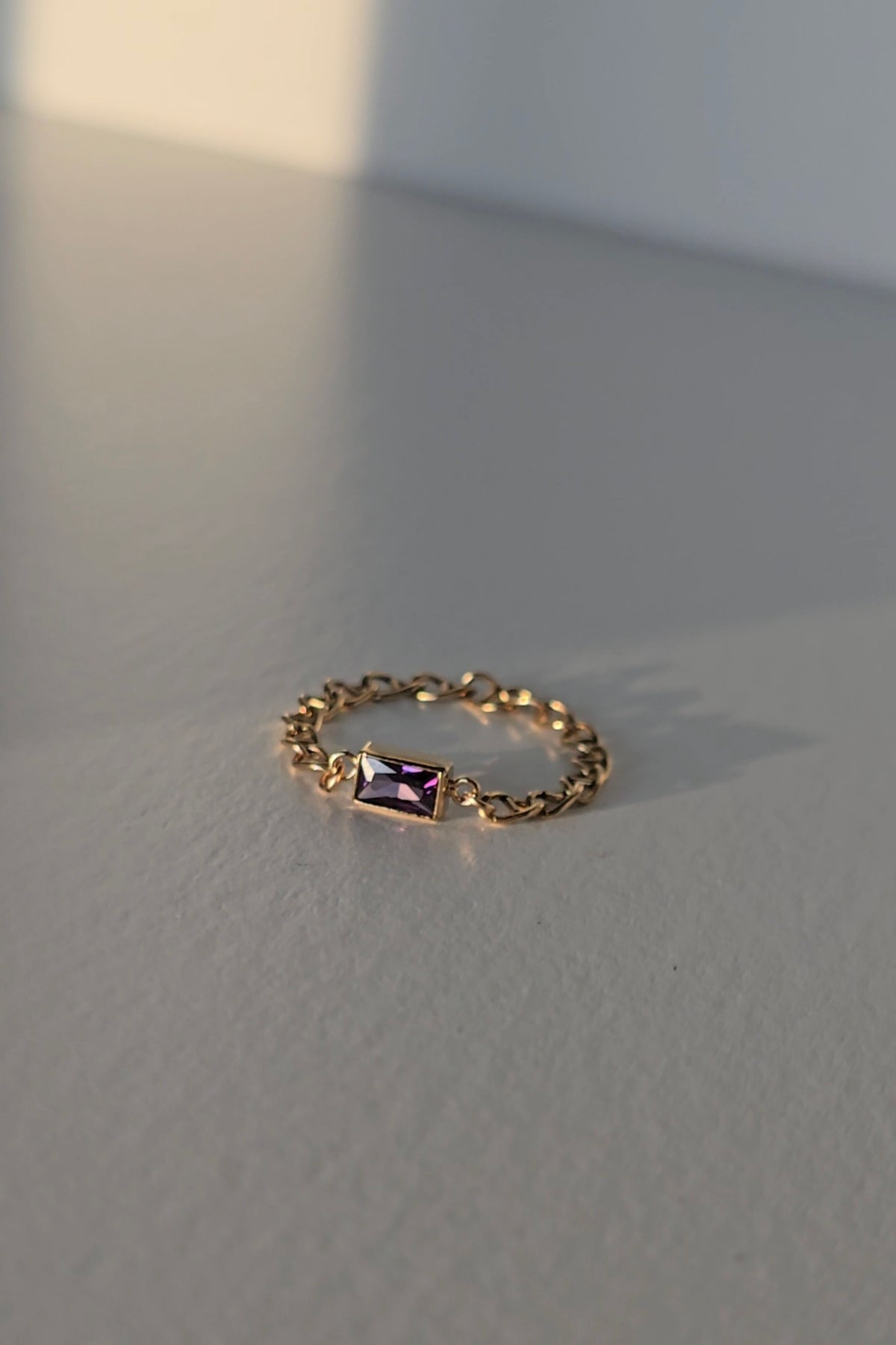 A delicate gold chain ring featuring a small rectangular purple crystal, resting on a textured gray surface with soft, natural shadows.