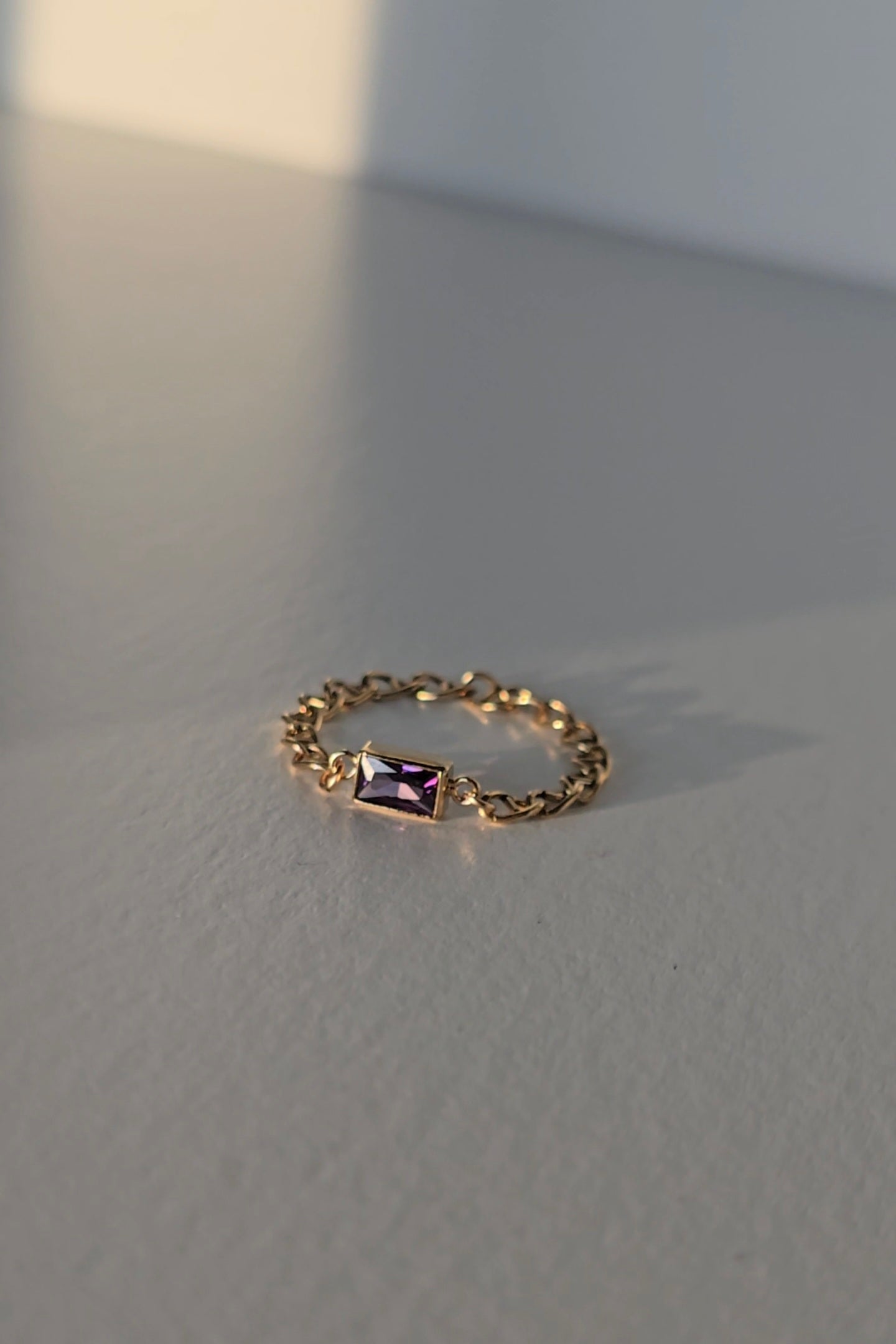 A delicate gold chain ring featuring a small rectangular purple crystal, resting on a textured gray surface with soft, natural shadows.
