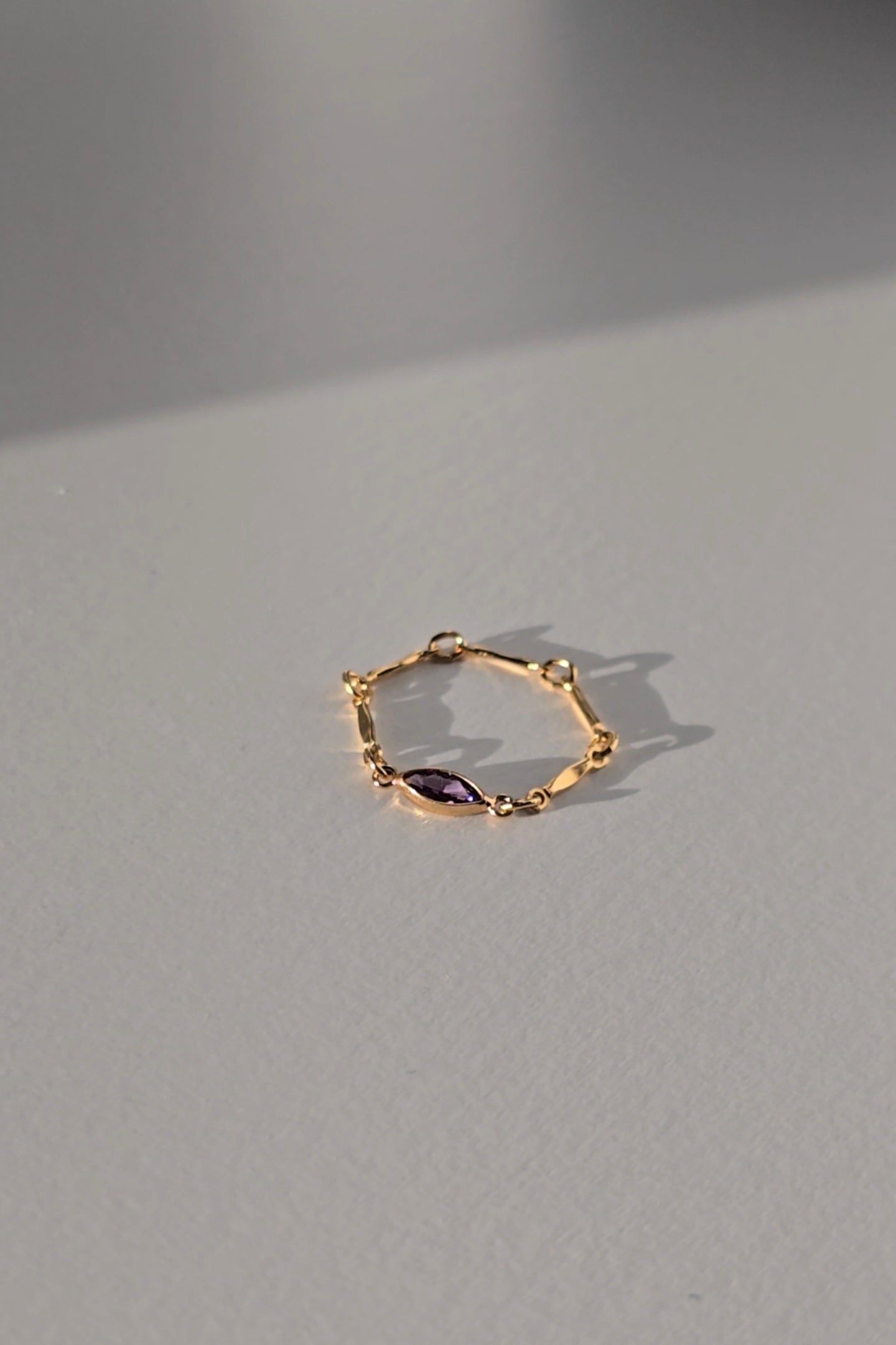 A delicate gold ring featuring a marquise-cut purple gemstone, resting on a light gray surface with soft shadows.