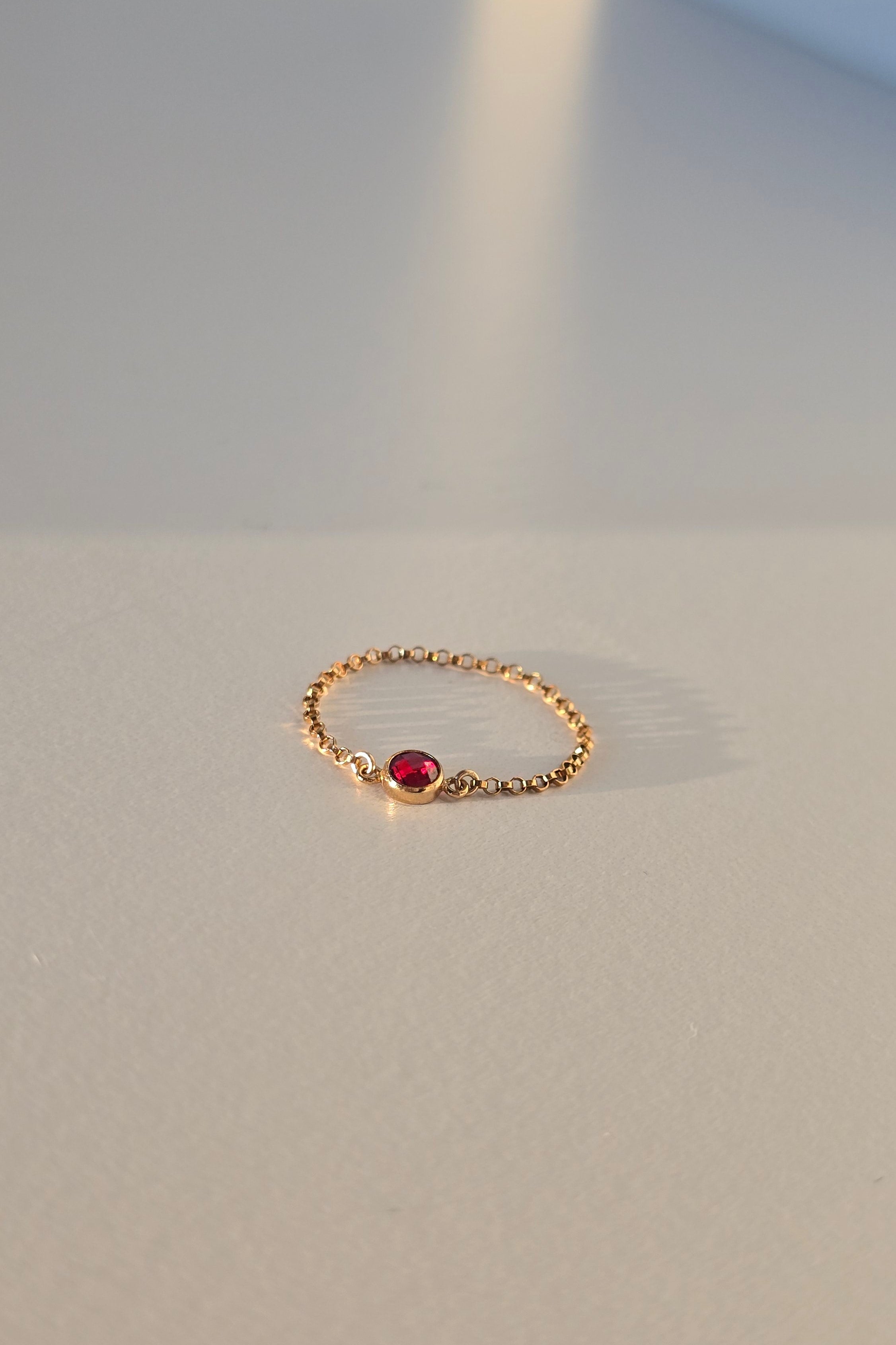 A delicate gold ring featuring a small, round red gemstone, displayed on a plain light gray wall.