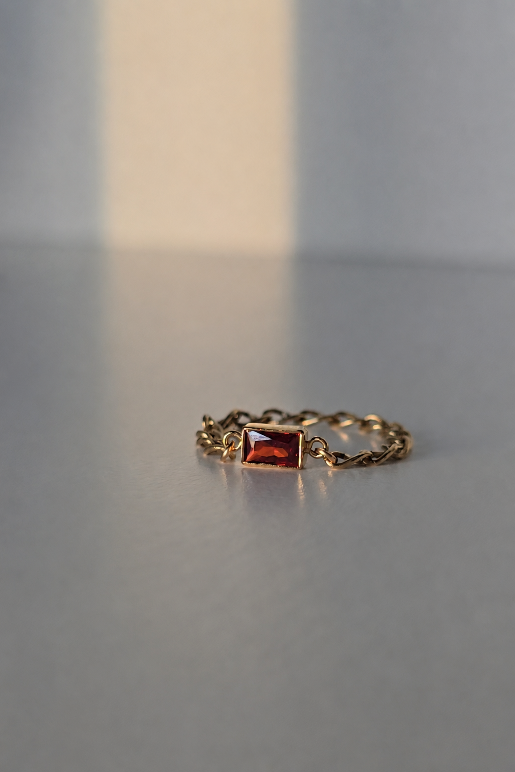 A delicate gold chain ring featuring a small rectangular red gemstone, resting on a smooth gray surface with soft, warm light casting a shadow.
