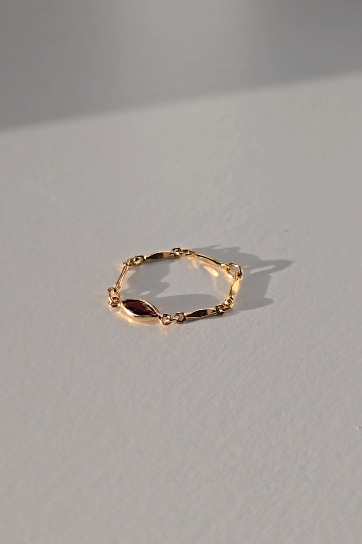 A delicate gold chain ring with a small polished gold marquise-shaped charm, resting on a light gray textured surface with a soft shadow.