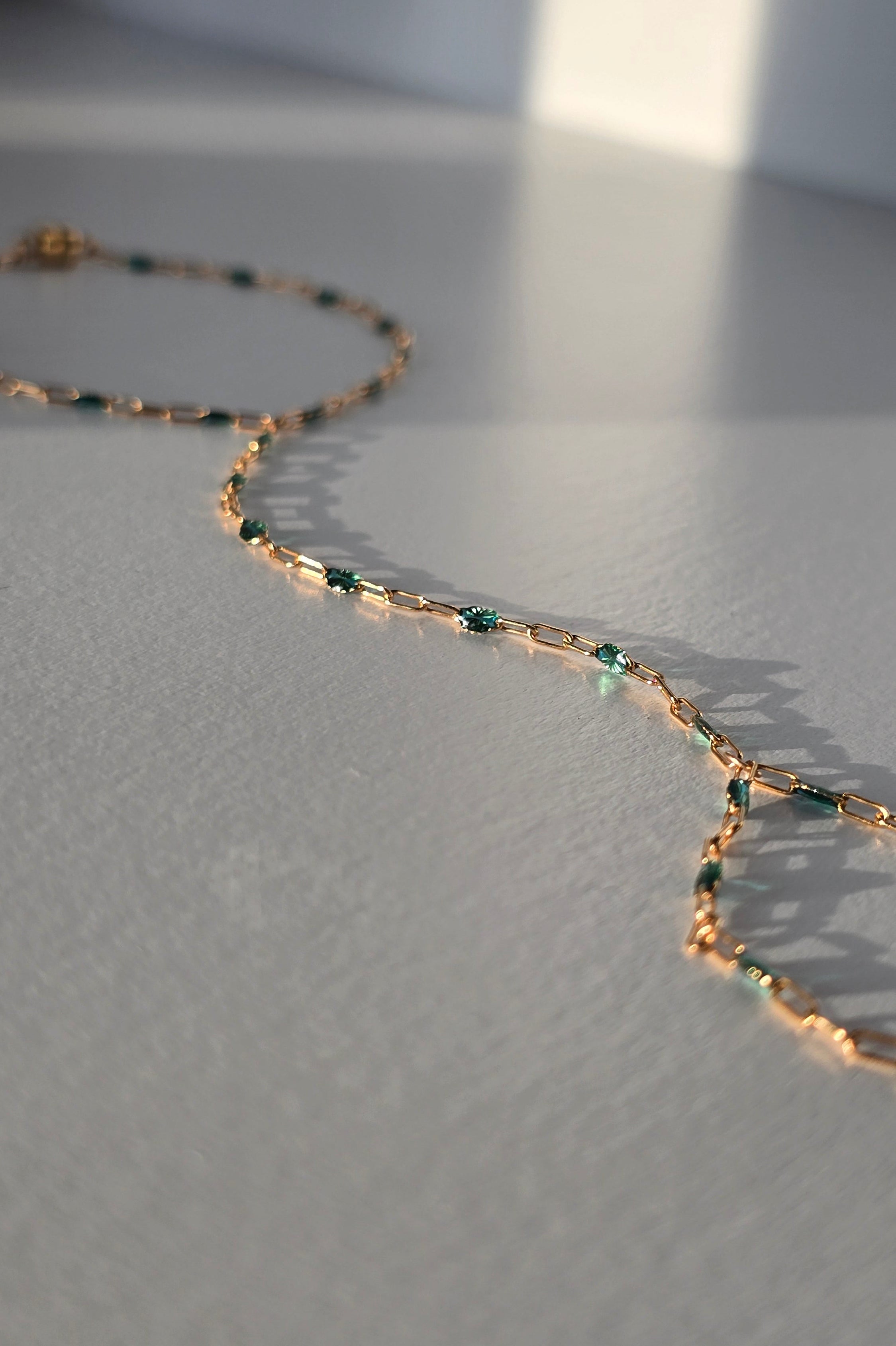A delicate gold hand chain with small teal gemstone accents, photographed on a light gray surface with soft natural shadows.