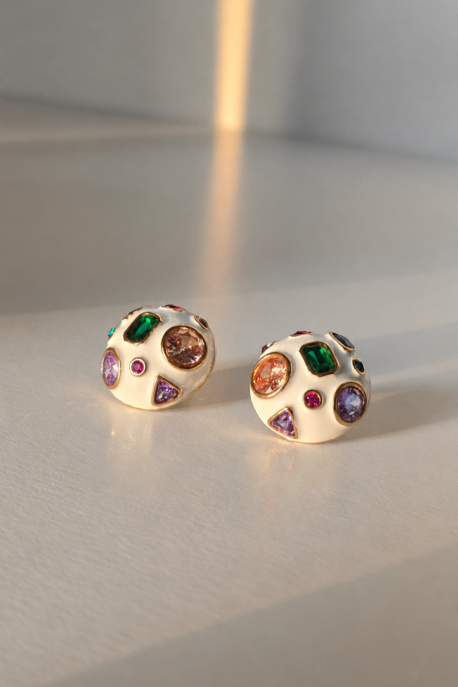 A pair of round gold-toned stud earrings with a cream-colored enamel face, decorated with multiple colorful gemstones in green, pink, purple, and orange, resting on a light gray surface.
