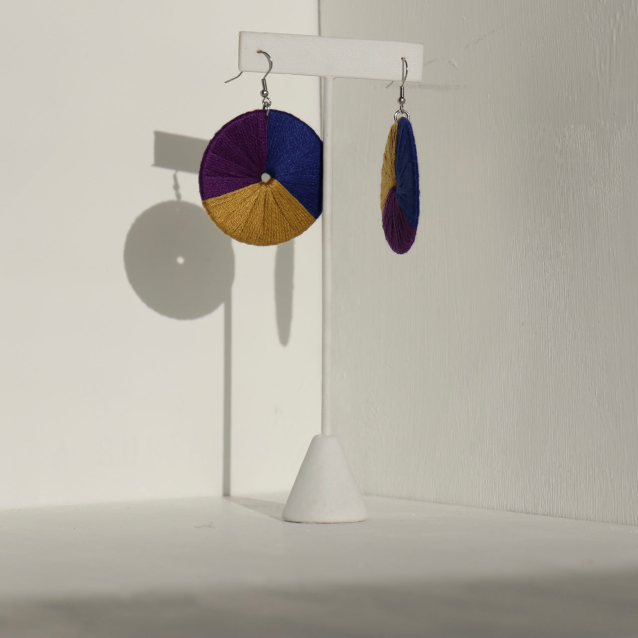 The Bode earrings in deep purple and gold, displayed on a stand with natural shadows