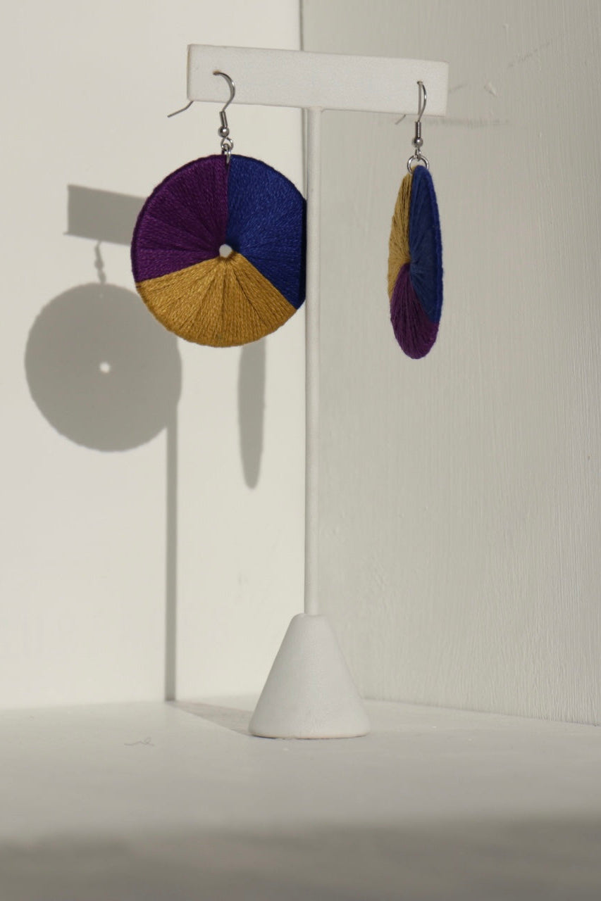 The Bode earrings in deep purple and gold, displayed on a stand with natural shadows