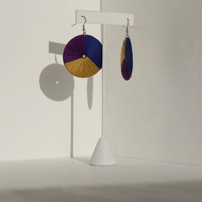 The Bode earrings in deep purple and gold, displayed on a stand with natural shadows